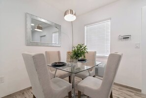 Een eetruimte met een glazen tafel, vier stoelen, een potplant en een spiegel aan de muur.