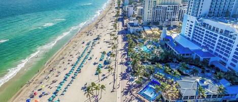 A beachside hotel with multiple levels, a pool area, and a beach with umbrellas.