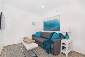 A living room with a grey sofa, teal cushions, a glass coffee table, a white side table with a lamp, and a painting of a wave on the wall. - Renovated studio w/ ocean views & boardwalk steps (Hollywood)