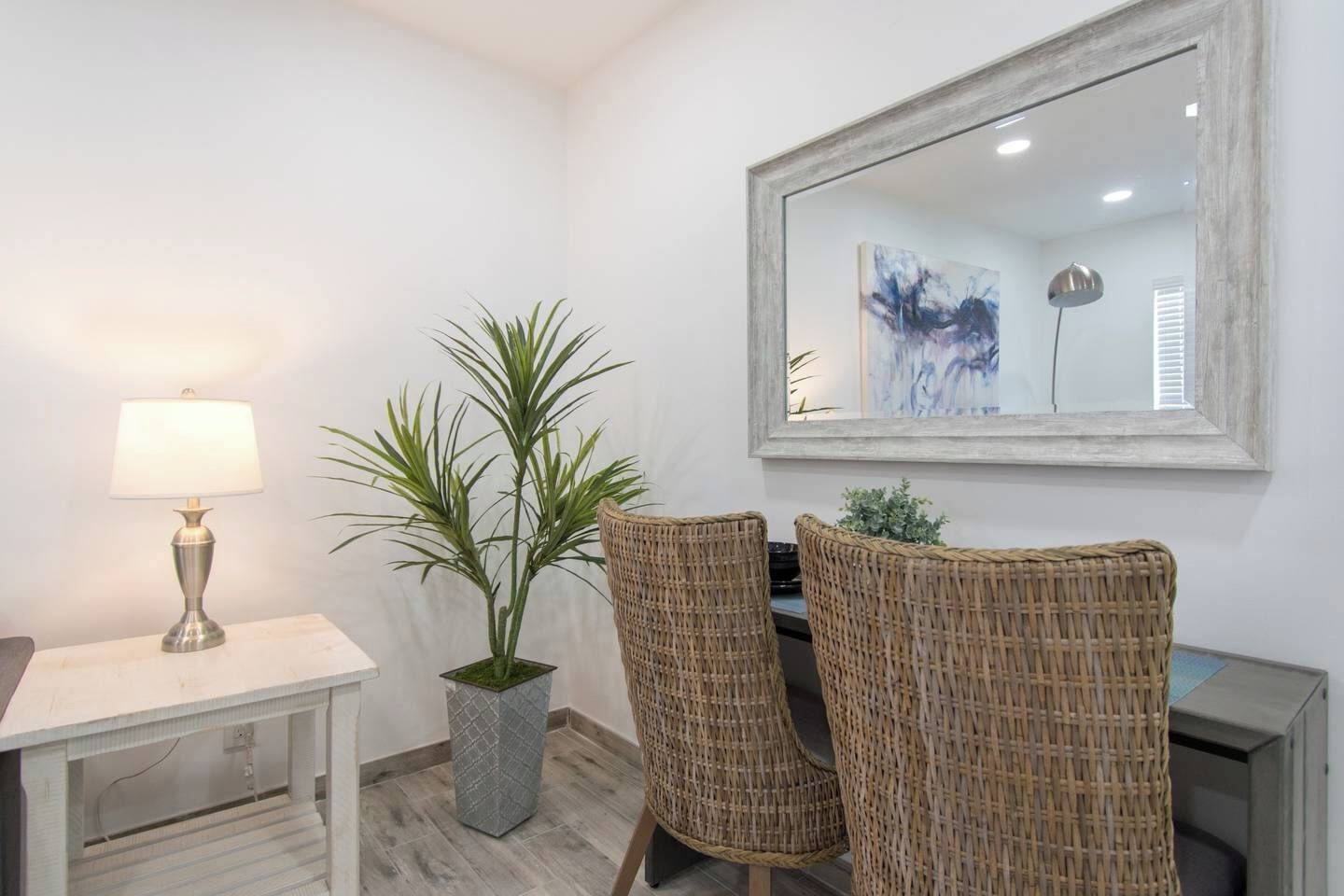 Una habitación con una mesa blanca, una lámpara, dos sillas de mimbre, una planta en maceta y un espejo grande en la pared.