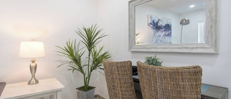 Una habitación con una mesa blanca, una lámpara, dos sillas de mimbre, una planta en maceta y un espejo grande en la pared.