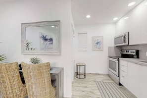 A modern kitchen with white cabinetry, a microwave, and an oven. There are two wicker chairs and a small round stool. - Modern apartment w/king bed & steps to beach (Hollywood)