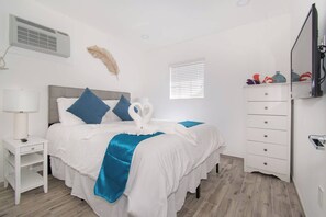 Une chambre d’hôtel comprenant un lit, une table de chevet, une commode, un téléviseur et une fenêtre avec des stores.