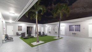 Een modern gebouw met een overdekt buitengebied met kunstgras, lounge stoelen en palmbomen.