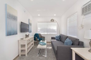 Ein modernes Wohnzimmer mit grauem Sofa, einem weißen Couchtisch, einem Schreibtisch mit Schubladen, einem Sessel und einem großen Fenster mit Jalousien.