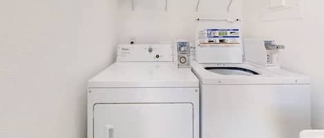 A laundry area with two front-loading washers and dryers, a metal shelf above, and a white wall.