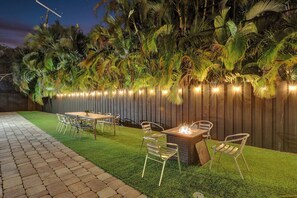 An outdoor patio with a wooden table and chairs, a fire pit, and a decorative fence with lights. - Family-Friendly Beach House, Steps to Beach (Fort Lauderdale)