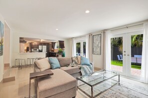 Een moderne woonkamer met een glazen salontafel, een bank met kussens, en een keuken op de achtergrond.