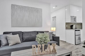 Ein modernes Wohnzimmer mit einem grauen Sofa, einem Couchtisch mit der Aufschrift 'RELAX' und einer schlichten Küche im Hintergrund.