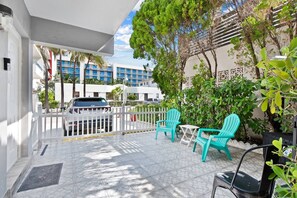 Eine Terrasse mit türkisfarbenen Stühlen und einem kleinen Tisch, ein weißer Zaun und ein Auto im Hintergrund.