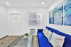 A modern living room with a blue sofa, a glass coffee table, and a large abstract painting on the wall. - Bright 1BR aparthotel near beach w/ free parking (Hollywood)