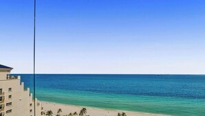 Blick auf einen Strand mit blauem Himmel, ruhiger See und einem Gebäude mit Balkonen.