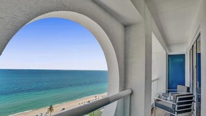 Ein Balkon mit Blick auf den Ozean und den Strand, eine Sitzecke mit Stühlen und ein Fenster mit Vorhängen.