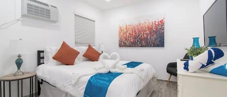 A hotel room with a bed, a desk, a chair, a lamp, a painting, and an air conditioning unit.