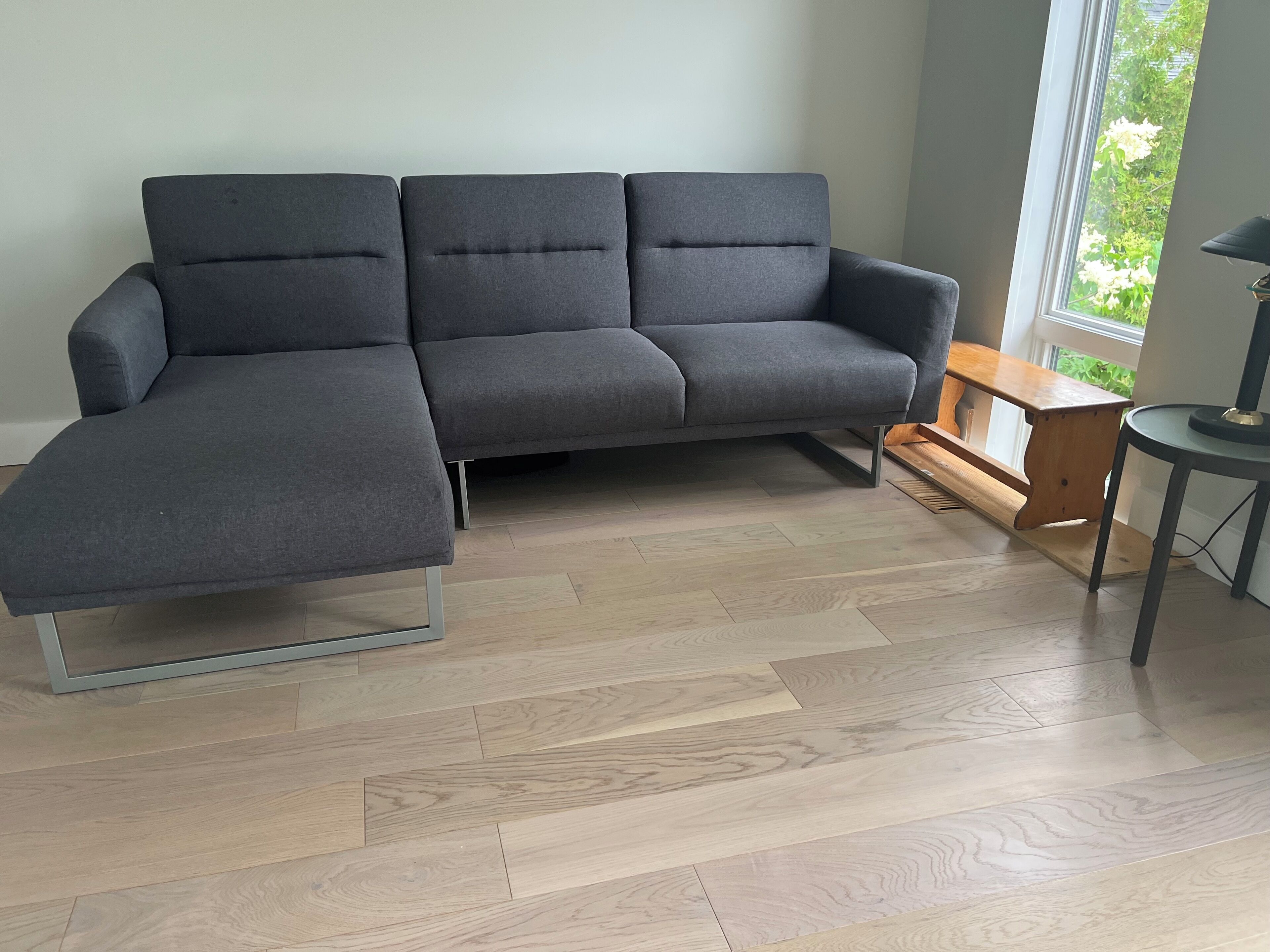 A grey sectional sofa with a metal frame, a wooden bench, and a small round table with a lamp.