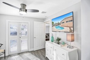A modern living room with a ceiling fan, a flat-screen TV mounted on the wall, a white console table with decorative items, and a door with blinds. - Spacious home w/pool, fire pit & grill (Lighthouse Point)