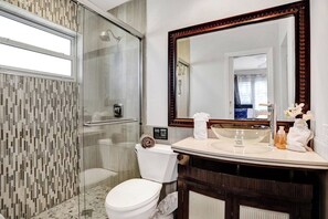 A modern bathroom with a glass shower enclosure, a white toilet, a vanity with a large mirror, and a sink. - Spacious home w/pool, fire pit & grill (Lighthouse Point)