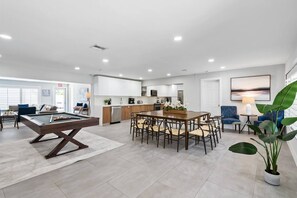 A spacious living area with a pool table, a dining table with chairs, a kitchen area, and a seating area with a lamp and a large plant. - Spacious home w/pool, fire pit & grill (Lighthouse Point)