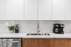 A modern kitchen with white cabinets, a stainless steel sink, a coffee maker, and a potted plant. - Spacious home w/pool, fire pit & grill (Lighthouse Point)