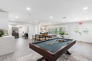 A modern living area with a pool table, a dining table with chairs, a kitchen with a refrigerator, and a large window with blinds. - Spacious home w/pool, fire pit & grill (Lighthouse Point)