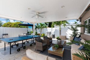 An outdoor patio with a ping pong table, seating area, and a ceiling fan. - Spacious home w/pool, fire pit & grill (Lighthouse Point)