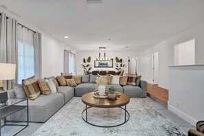 A modern living room with a grey sectional sofa, a round coffee table, and a dining area in the background. - Family villa w/ heated pool, gameroom & fire pit (Davie)