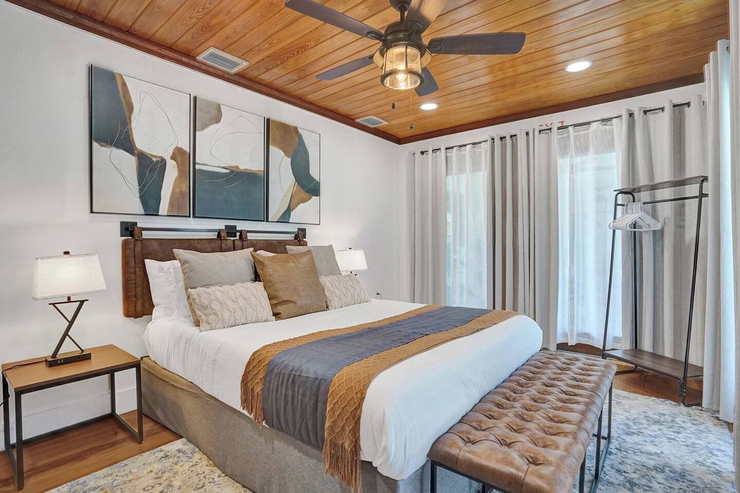 A bedroom with a bed, bedside tables, a ceiling fan, artwork, curtains, and a bench.
