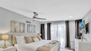 Une chambre à coucher moderne avec un lit, un ventilateur de plafond, des œuvres d’art accrochées au mur et un téléviseur sur le côté.