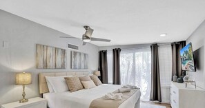 Une chambre à coucher moderne avec un lit, un ventilateur de plafond, des œuvres d’art accrochées au mur et un téléviseur sur le côté.