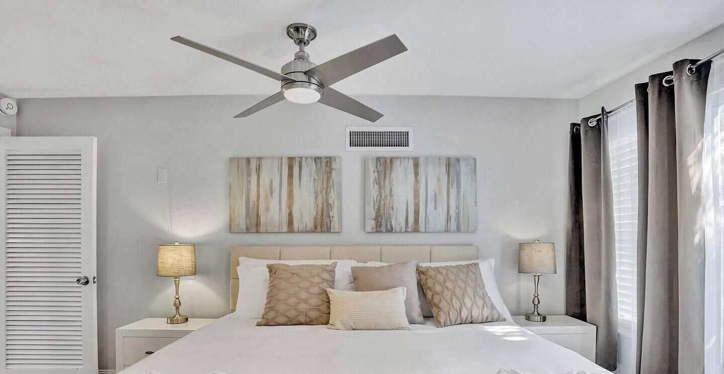 A hotel room with a bed, two bedside tables, a ceiling fan, artwork on the wall, and curtains.