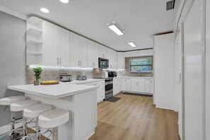 A modern kitchen with white cabinets, a central island with bar stools, a microwave, and a window with a view. - Modern house w/ heated pool & game room (Pompano Beach)