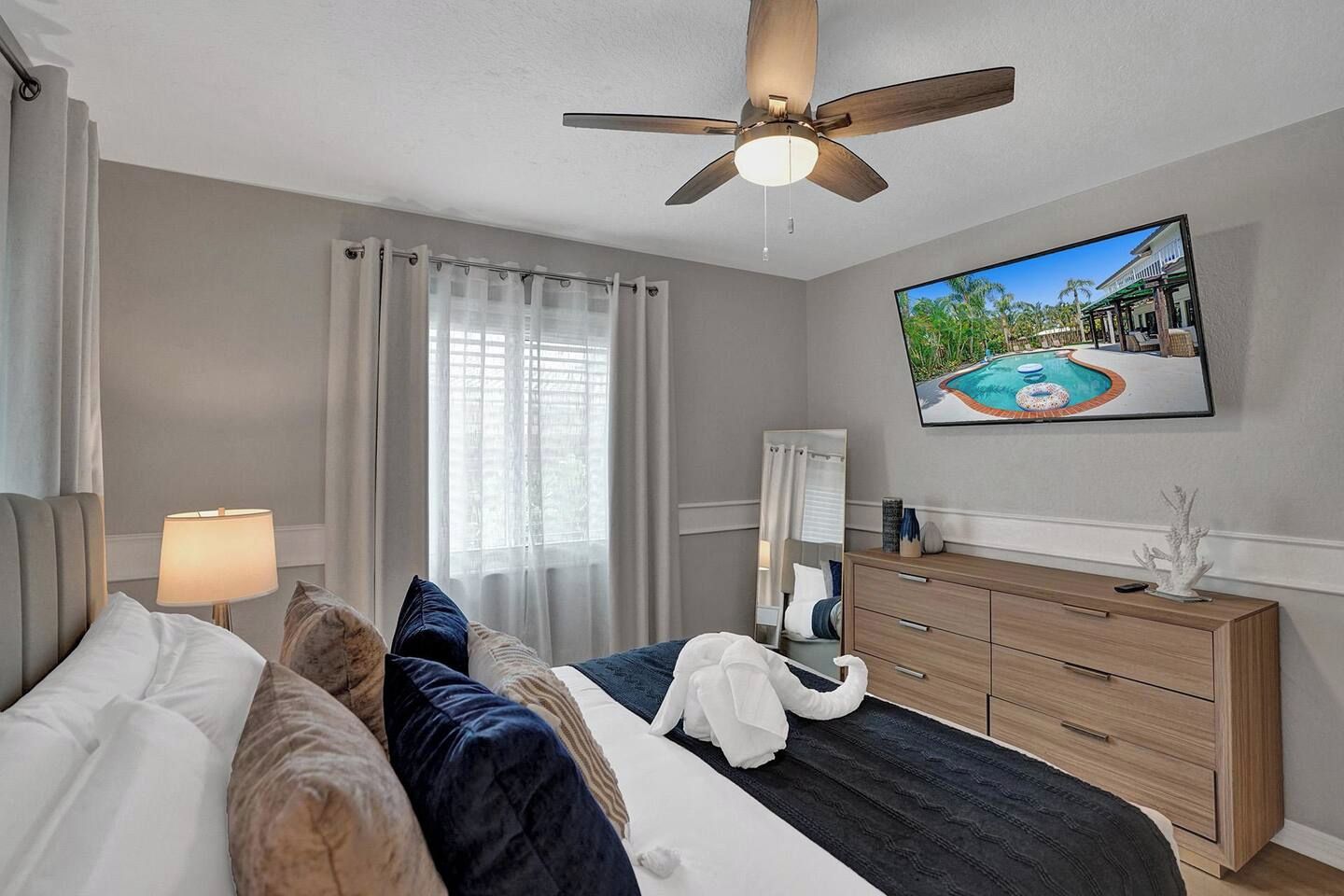 A hotel room with a bed, a ceiling fan, a TV displaying a pool, a wooden dresser, and a window with curtains.