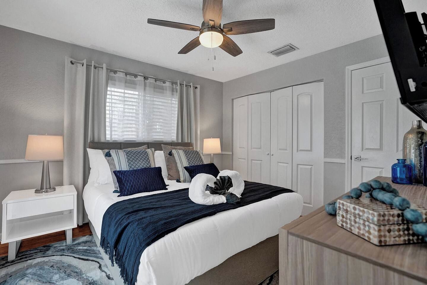 Une chambre à coucher comprenant un lit, des tables de chevet, un ventilateur de plafond, une fenêtre avec des rideaux et une commode agrémentée d’objets décoratifs.