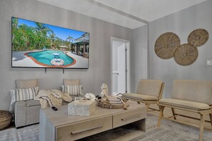 A living room with a television displaying a pool scene, a beige sofa, a wooden coffee table, and a door. - Modern house w/ heated pool & game room (Pompano Beach)