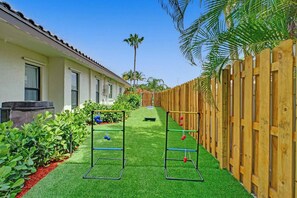 Un patio con césped artificial, dos estructuras de equipo de gimnasia metálicas, una valla de madera y un edificio con ventanas.