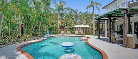 A swimming pool with a wooden deck, surrounded by palm trees and a covered seating area.