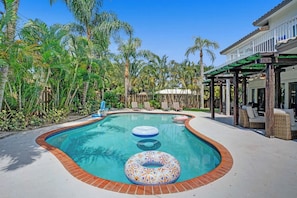 A swimming pool with a wooden deck, surrounded by palm trees and a covered seating area. - Modern house w/ heated pool & game room (Pompano Beach)