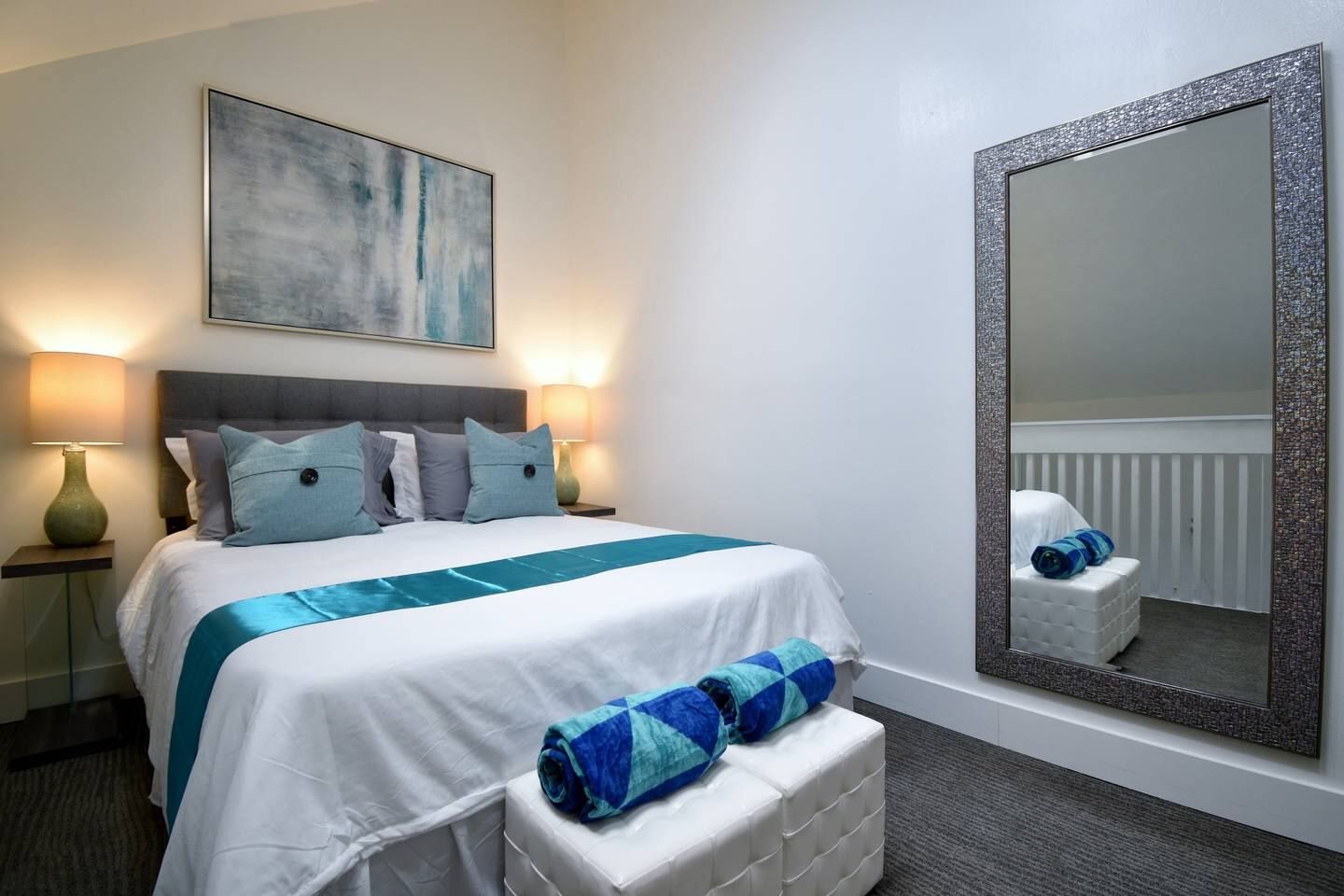 A hotel room with a bed, bedside lamps, a large mirror, and a painting on the wall.