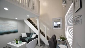 Ein modernes Wohnzimmer mit Treppe, einem Essbereich und einer durch ein Fenster sichtbaren Küche.