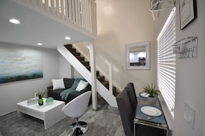 Ein modernes Wohnzimmer mit Treppe, einem Essbereich und einer durch ein Fenster sichtbaren Küche.