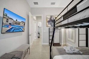 Habitación de hotel con literas, televisor de pantalla plana, un banco y un pasillo que conduce a un baño.
