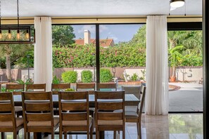 A dining area with wooden chairs and tables, large windows with curtains, and a view of a garden and pool. - Pool - Soccer - Movie Room - Game Room - Sleeps 20 (Largo)