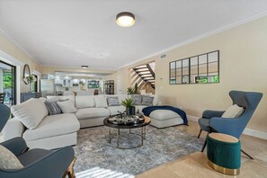 Amplia sala de estar con un sofá modular, una mesa de centro y un sillón azul. Se observa una cocina al fondo.