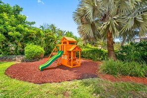 Een speeltuin met een glijbaan en een klein huisje, omringd door een tuin met diverse planten en een palmboom.