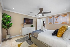 A bedroom with a bed, a ceiling fan, a TV, a dresser, a plant, and artwork on the wall.