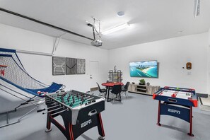 A room with two foosball tables, a projector screen, and a television displaying a scenic view. - Family estate w/private pool & basketball court