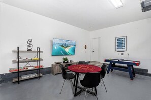 Et værelse med et fjernsyn monteret på væggen, et billardbord, et rødt bord med stole og en reol med diverse genstande.