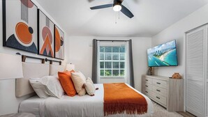 Une chambre à coucher comprenant un lit, un téléviseur, un ventilateur de plafond et des œuvres d’art accrochées au mur.