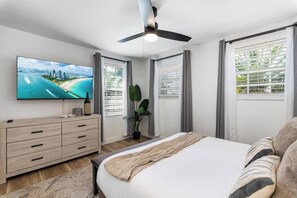 Un dormitorio moderno con cama, televisor, ventilador de techo y vista a una playa por la ventana.