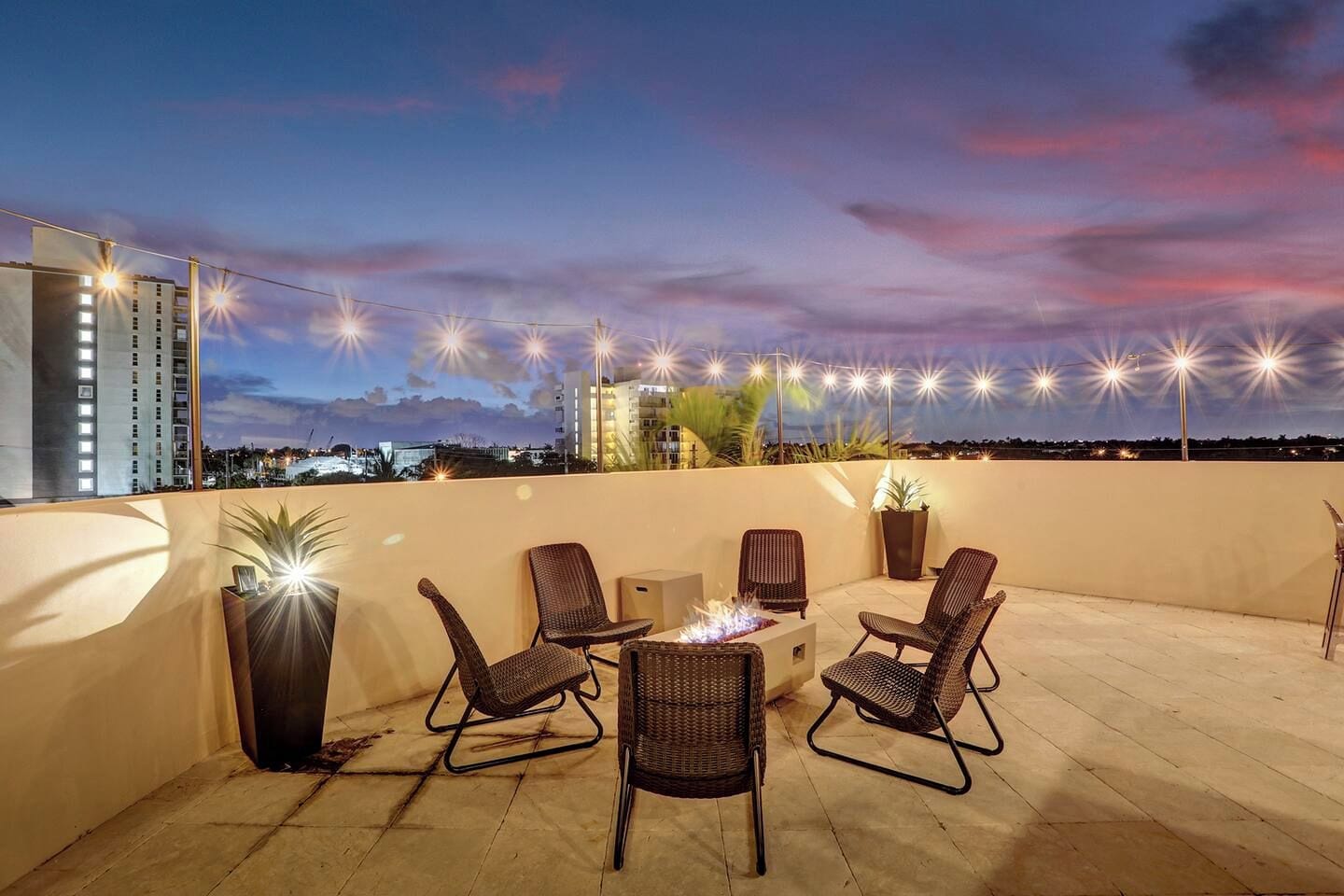A rooftop terrace with outdoor seating, a fire pit, string lights, and a cityscape view.